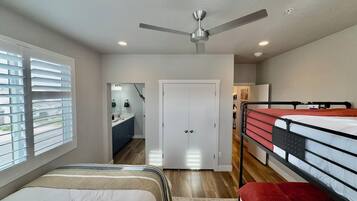 Une chambre avec des lits superposés, un ventilateur de plafond, une salle de bain avec un miroir et un lavabo, et une porte donnant sur une autre pièce.