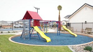 A playground with slides and a climbing structure, surrounded by a fence and a building in the background.