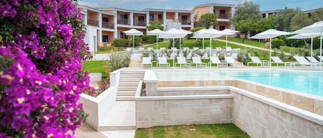 Un espace piscine doté de chaises longues et de parasols, entouré d’une pelouse bien entretenue et d’un bâtiment avec des balcons.