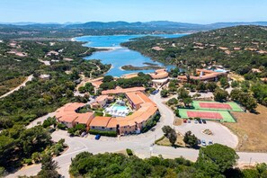 Vue aérienne d’une station balnéaire comprenant plusieurs bâtiments, une piscine, des courts de tennis et une vaste étendue d’eau.