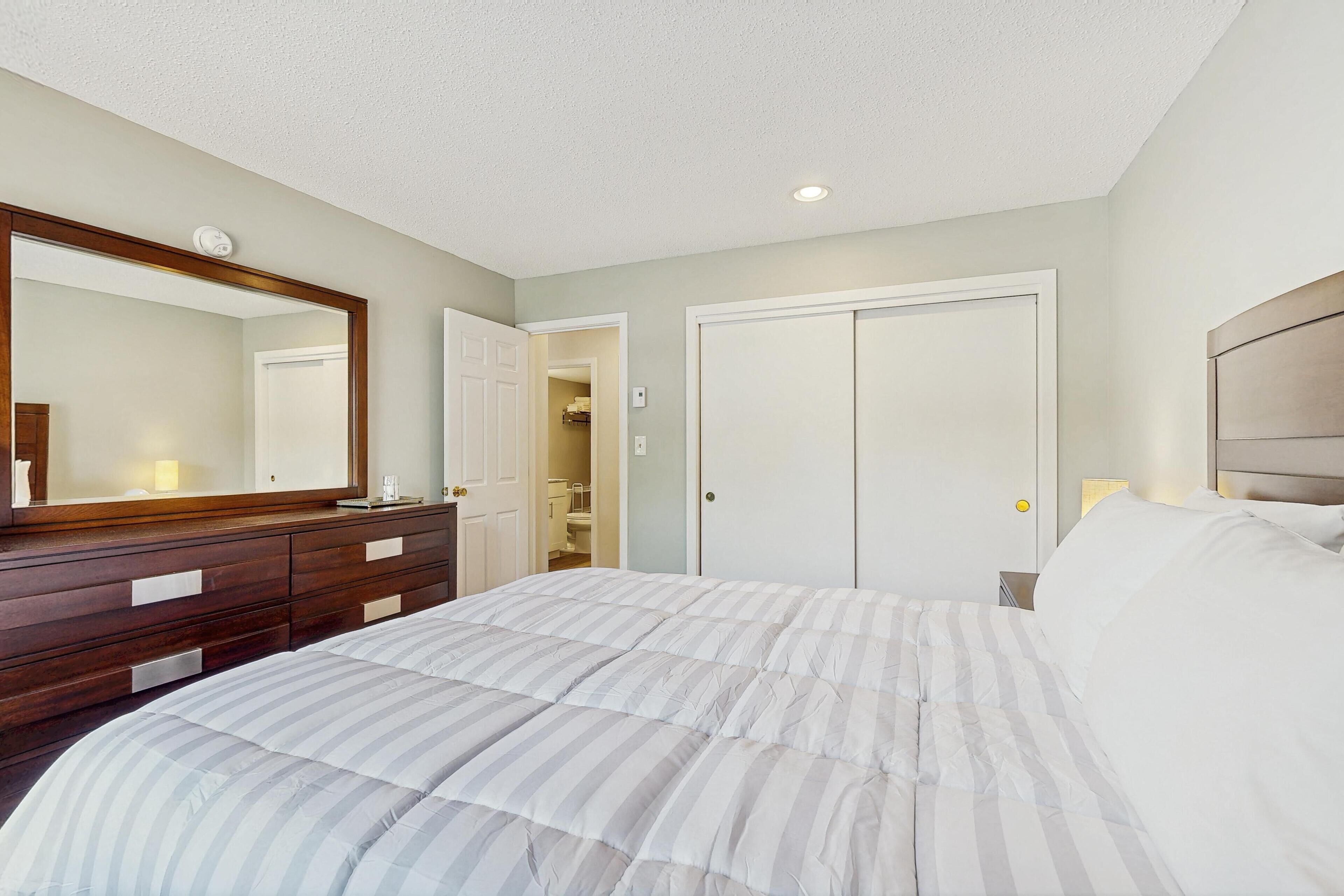 Une chambre à coucher comprenant un lit, une commode, un miroir, un placard et une porte donnant sur une salle de bain.