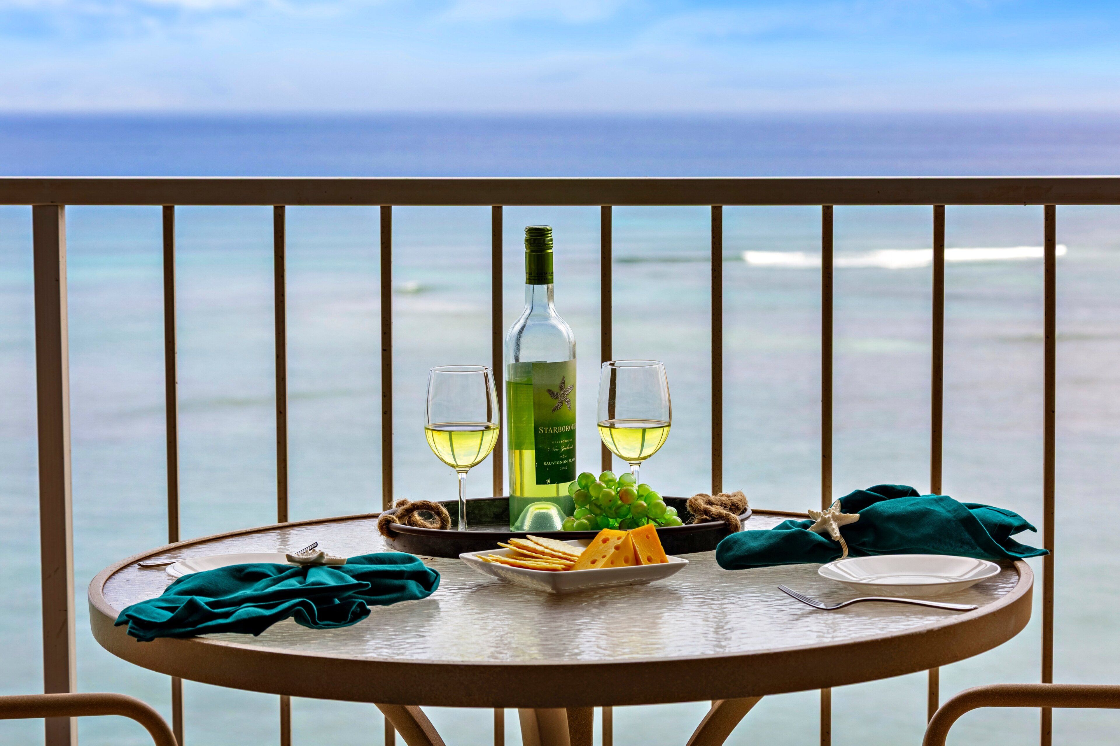 A table with a bottle of wine, two glasses, a plate of cheese, grapes, and a starfish on a balcony with a view of the ocean.