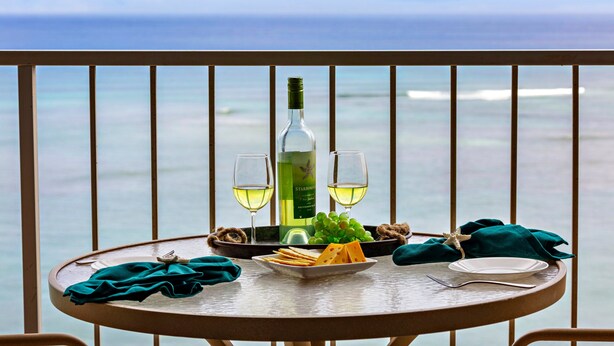 A table with a bottle of wine, two glasses, a plate of cheese, grapes, and a starfish on a balcony with a view of the ocean. - High-Rise Paradise with Sweeping Ocean Views (Lahaina)