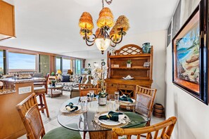 A dining area with a round glass table, wicker chairs, and a chandelier. - High-Rise Paradise with Sweeping Ocean Views (Lahaina)