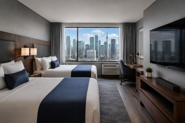 A hotel room with two beds, a desk, a TV, and a large window with a city view. - Modern Apartments (Toronto)