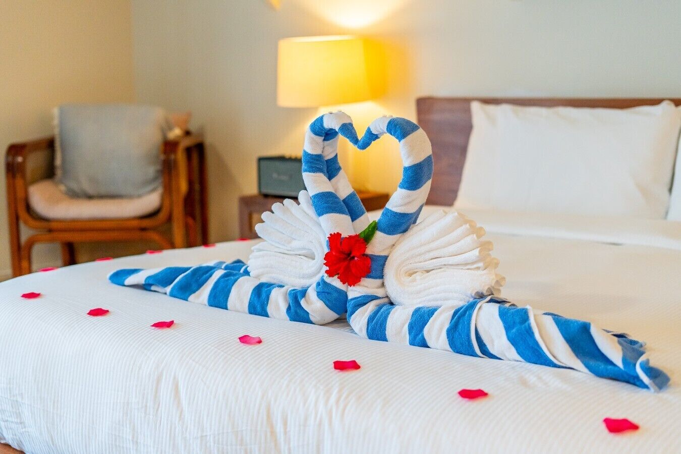 Een net opgemaakt bed, met een handdoek gevouwen in de vorm van een hart, een rode bloem en rozenblaadjes eromheen.