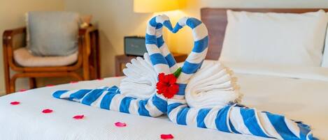 Een net opgemaakt bed, met een handdoek gevouwen in de vorm van een hart, een rode bloem en rozenblaadjes eromheen.