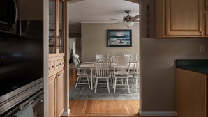 Uma cozinha com armários de madeira, uma mesa de jantar com cadeiras, um ventilador de teto e um quadro na parede.