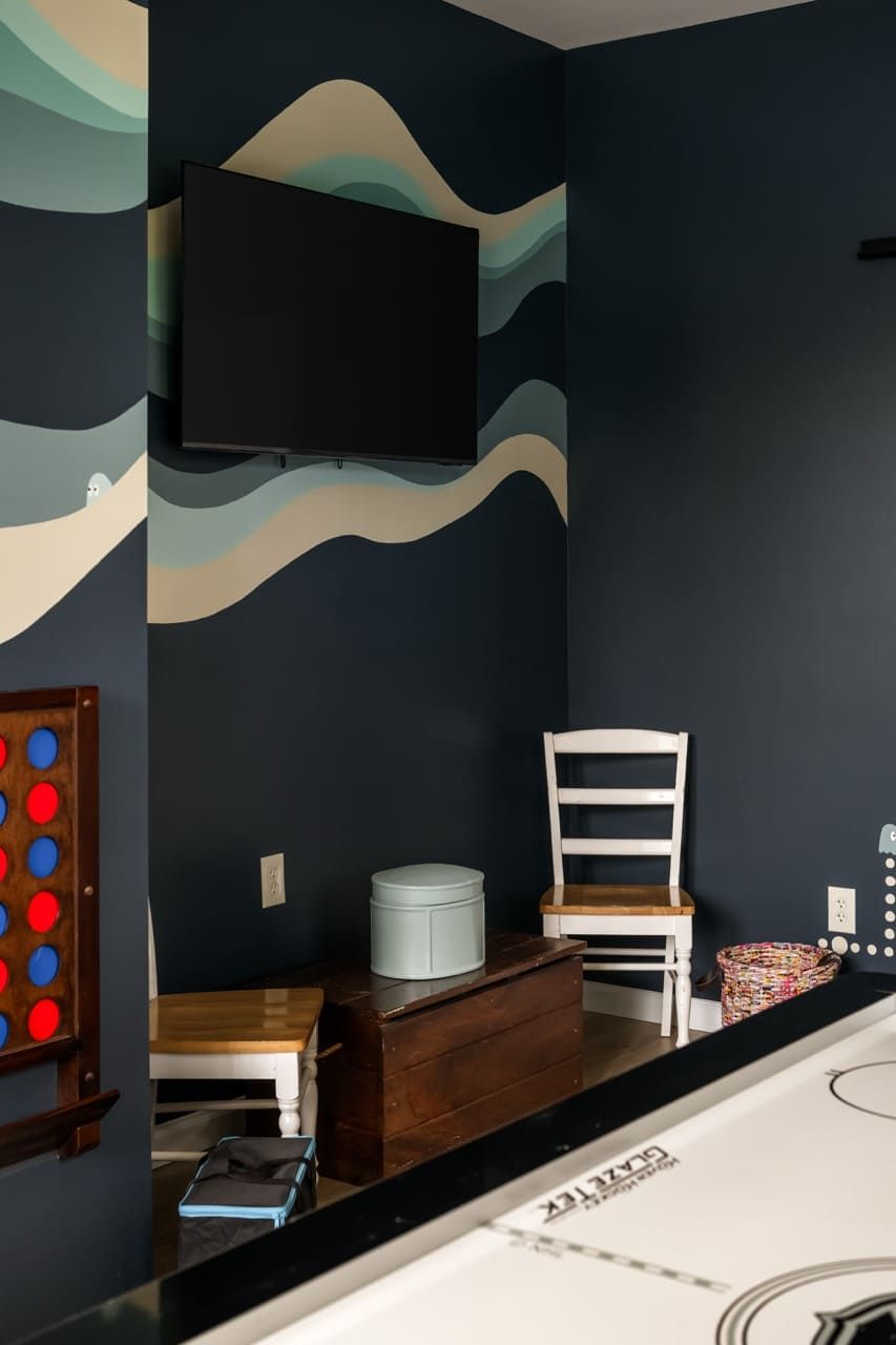A room with a dark wall, a mounted television, a wooden chair, a small table, and a white trash bin.