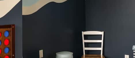 A room with a dark wall, a mounted television, a wooden chair, a small table, and a white trash bin.