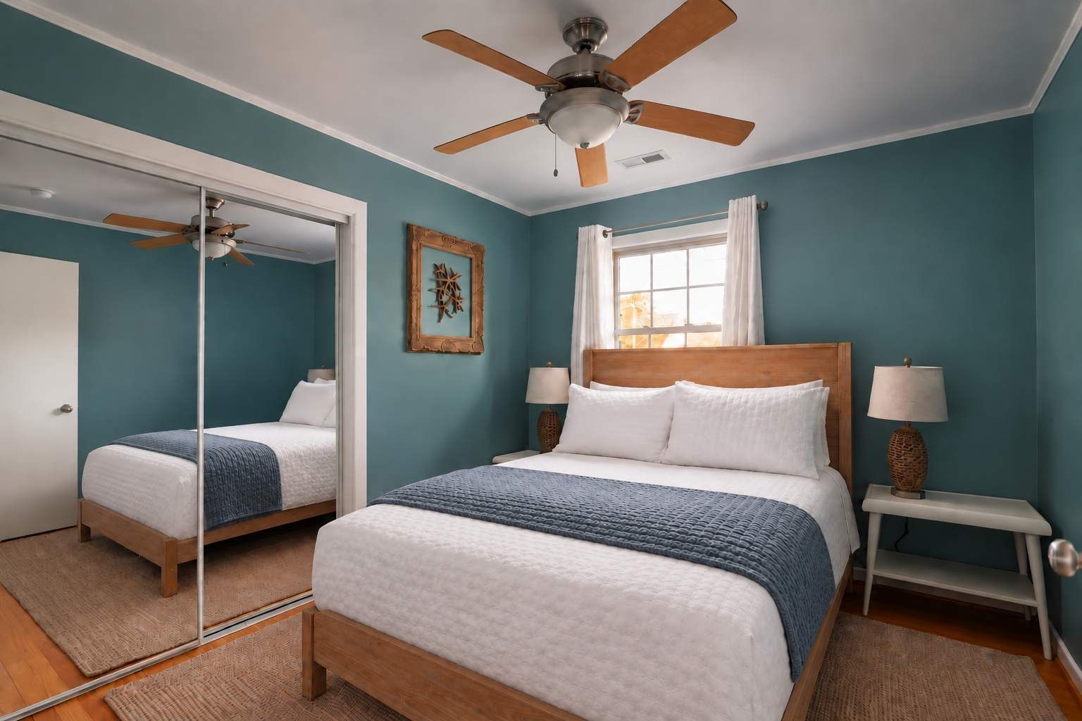 A bedroom with a bed, two bedside tables, a ceiling fan, a window with curtains, and a mirror.