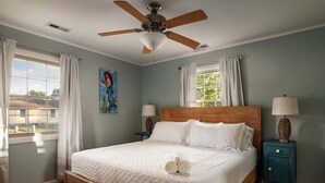 Quarto com cama, ventilador de teto, duas mesas de cabeceira, janela com cortinas e um quadro na parede.