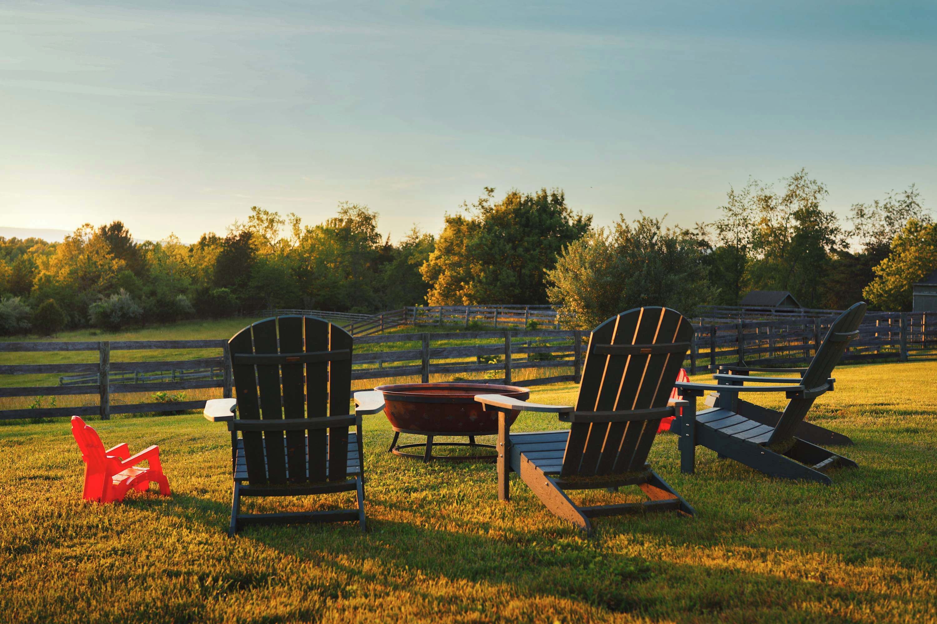 Zwei Adirondack-Stühle, ein roter Adirondack-Stuhl und ein Holztisch in einer grünen Außenanlage mit Holzzaun und Bäumen im Hintergrund.