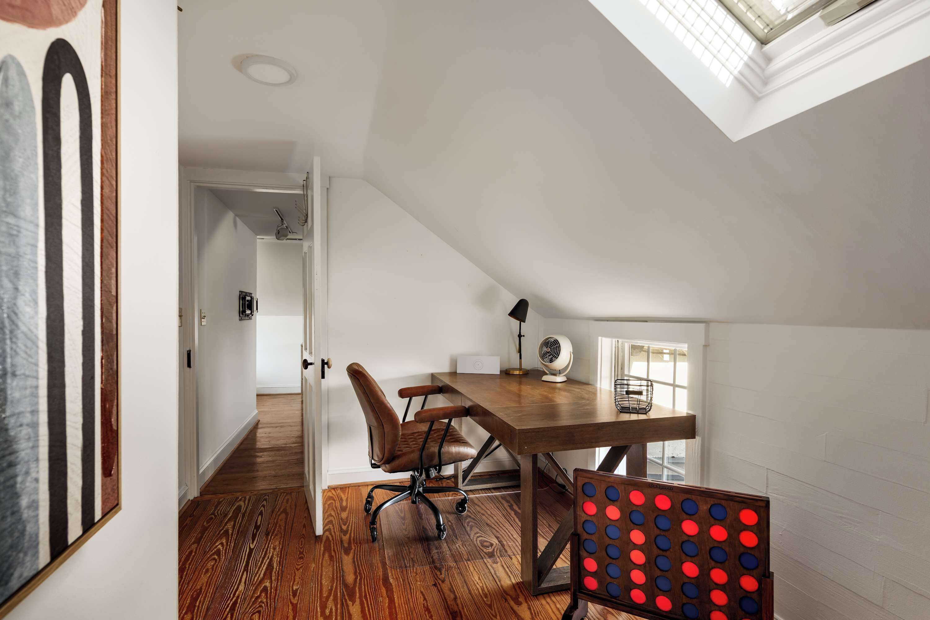 A room with a wooden floor, a desk with a chair, a ceiling fan, and a window.