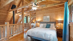 A wooden cabin bedroom with a bed, nightstands, a ceiling fan, and a staircase.