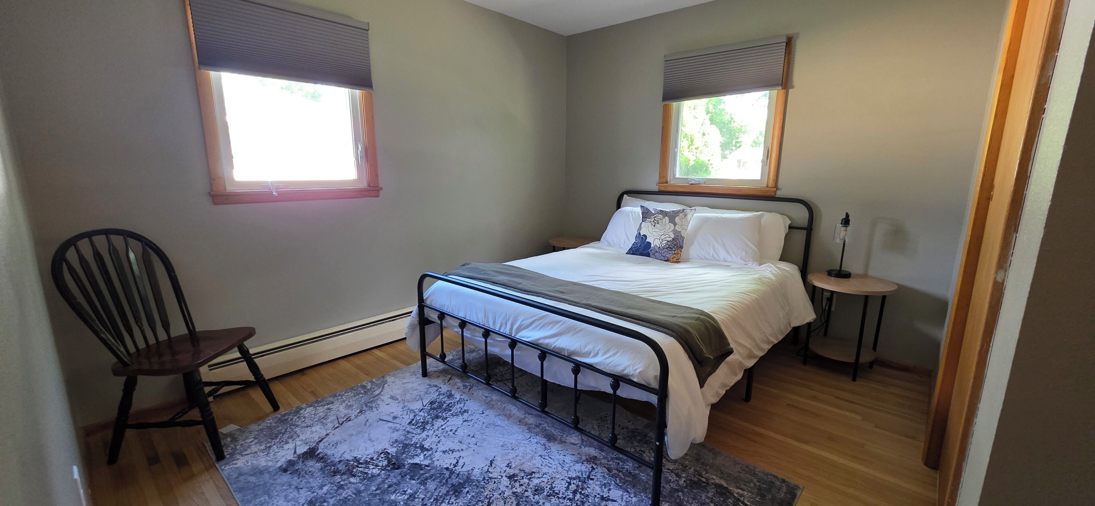 Une chambre à coucher comprenant un lit, une chaise, une table de chevet et deux fenêtres équipées de stores.