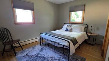 Une chambre à coucher comprenant un lit, une chaise, une table de chevet et deux fenêtres équipées de stores.