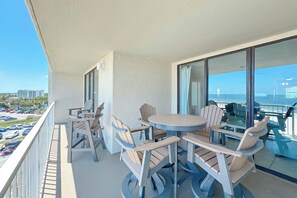 Een balkon met een ronde tafel en stoelen, grote ramen en uitzicht op een parkeerplaats en gebouwen.