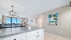 Une cuisine dotée d’un plan de travail en granit, de meubles blancs, d’un ventilateur de plafond et d’un tableau représentant une scène de plage.