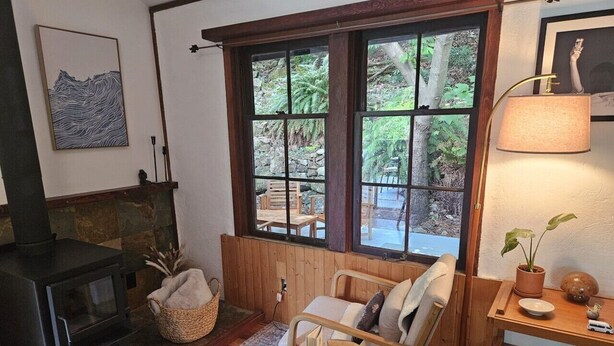Ein gemütliches Zimmer mit Kaminofen, einem bequemen Sessel, einem kleinen Tisch mit Lampe und Blick durch große Fenster.