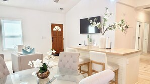 Ein modernes Wohnzimmer mit einem Glastisch, gepolsterten Sesseln, einem Holztisch, einem Flachbildfernseher und einer Zierpflanze.