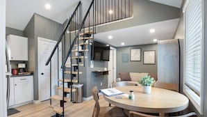 A modern kitchen with a wooden staircase, a dining area with a round table, and a living space with a sofa and a window.