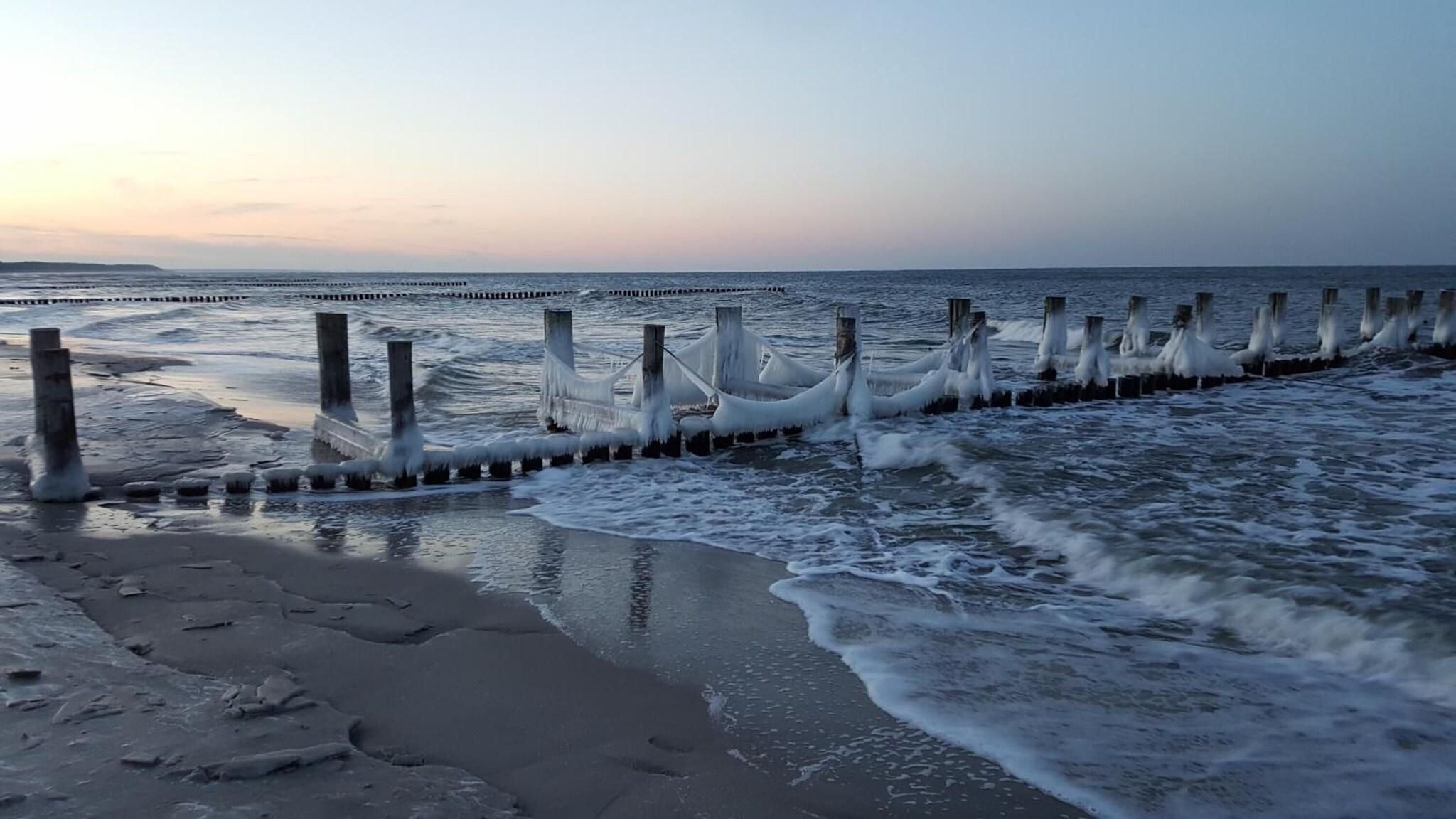 Ein Strand mit brechenden Wellen, die auf eine Reihe Holzpfähle treffen.