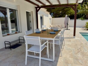 Ett takförsett utomhusområde med ett träbord och vita stolar, en pool i bakgrunden och en pergola.