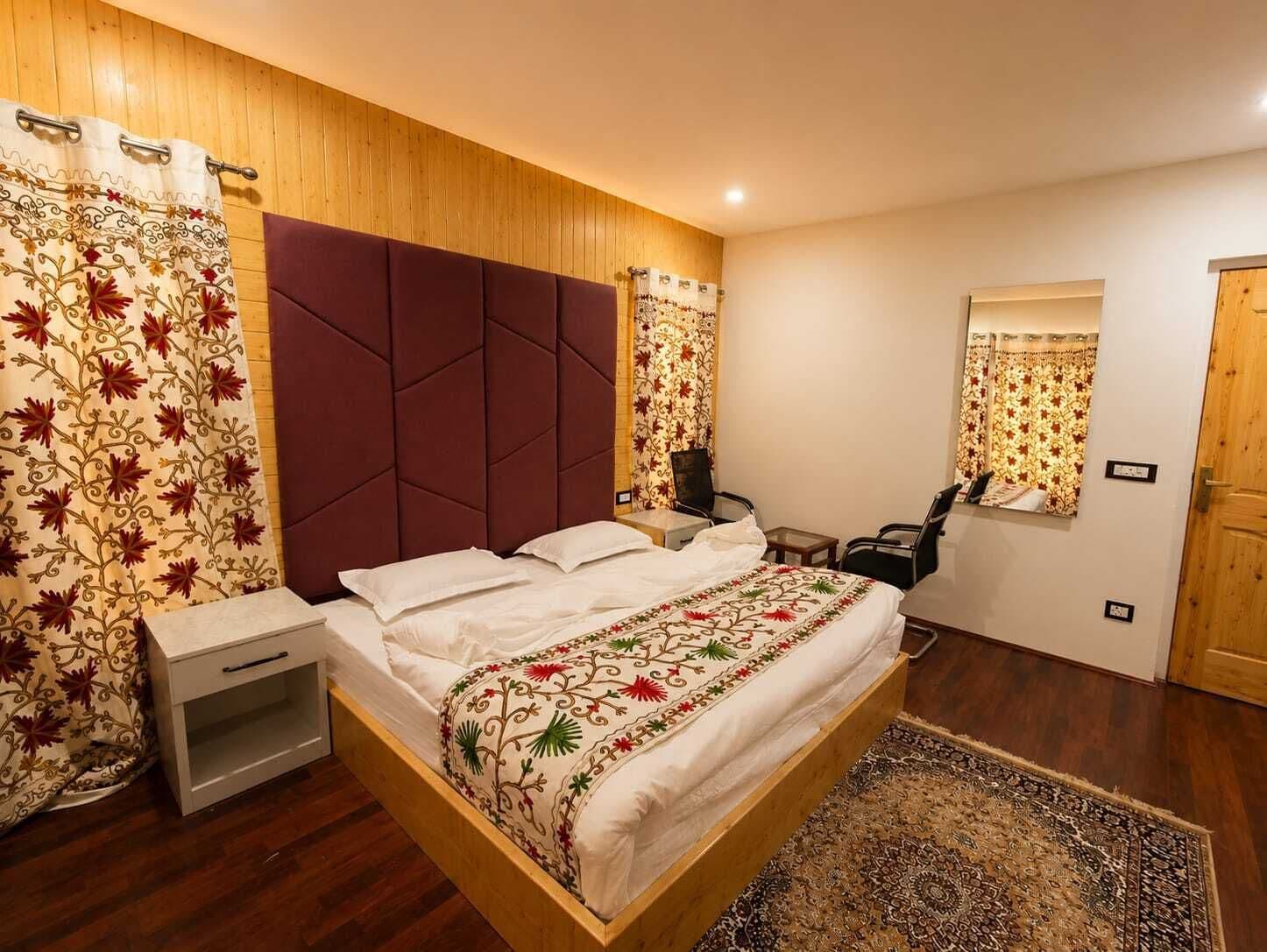 A bedroom with a wooden headboard, a bed with a floral bedspread, a nightstand, a chair, a small table, a mirror, and a patterned rug.