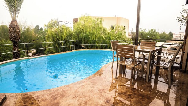 A swimming pool with a surrounding deck, a table with chairs, and a palm tree. - rose garden resort marrakech (Marrakech)