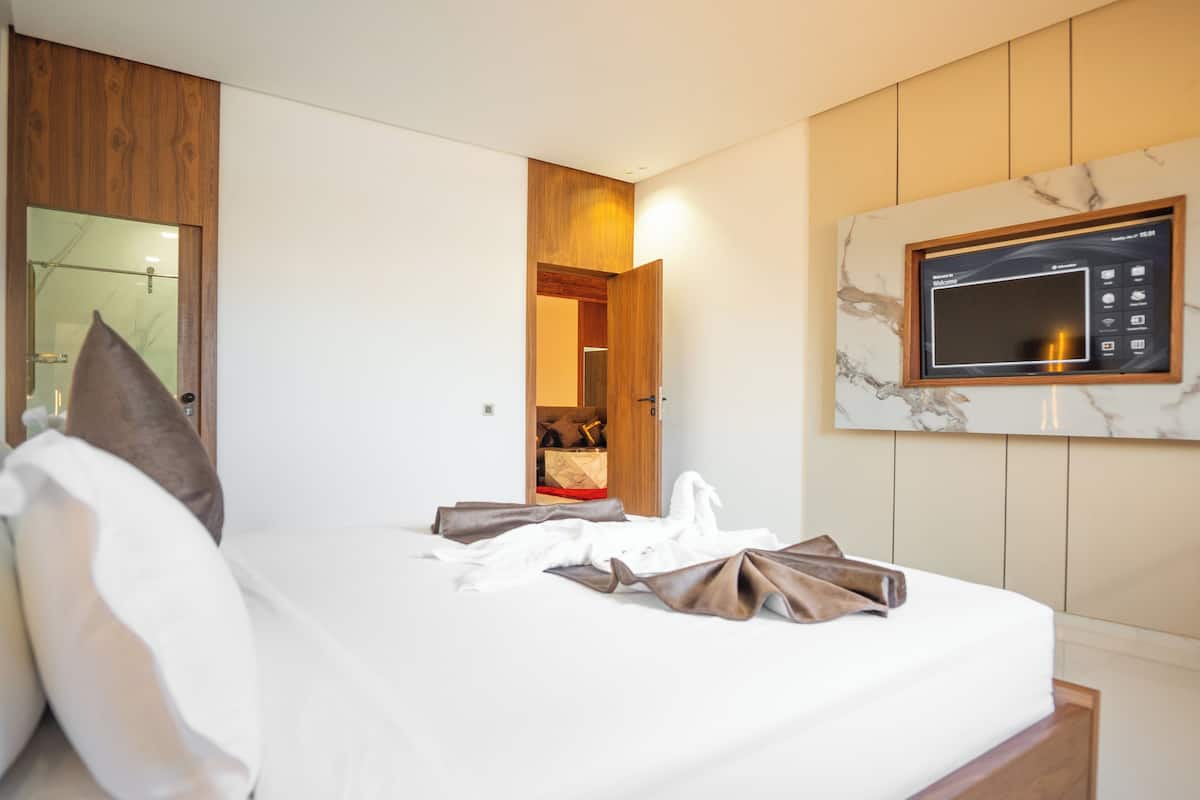 A neatly made bed with pillows, a wooden headboard, a wall-mounted TV, and a wooden door leading to a bathroom.