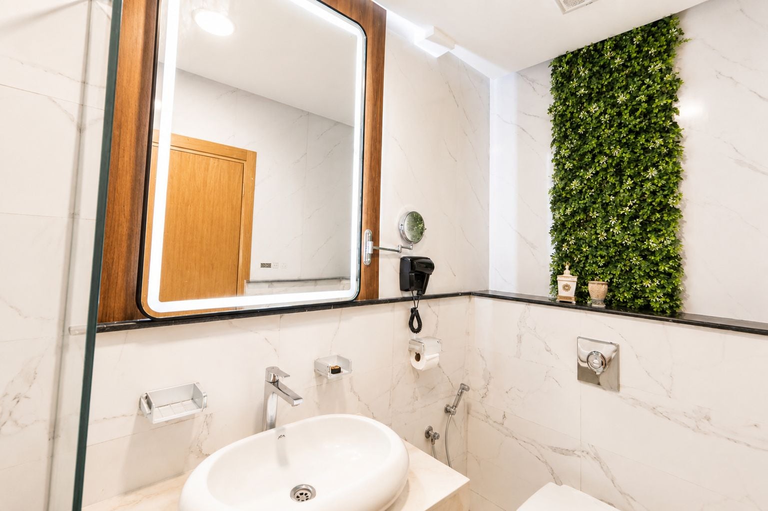 Un baño moderno con un lavabo de mármol, un espejo con marco de madera, un secador de pelo y un adorno verde en la pared.
