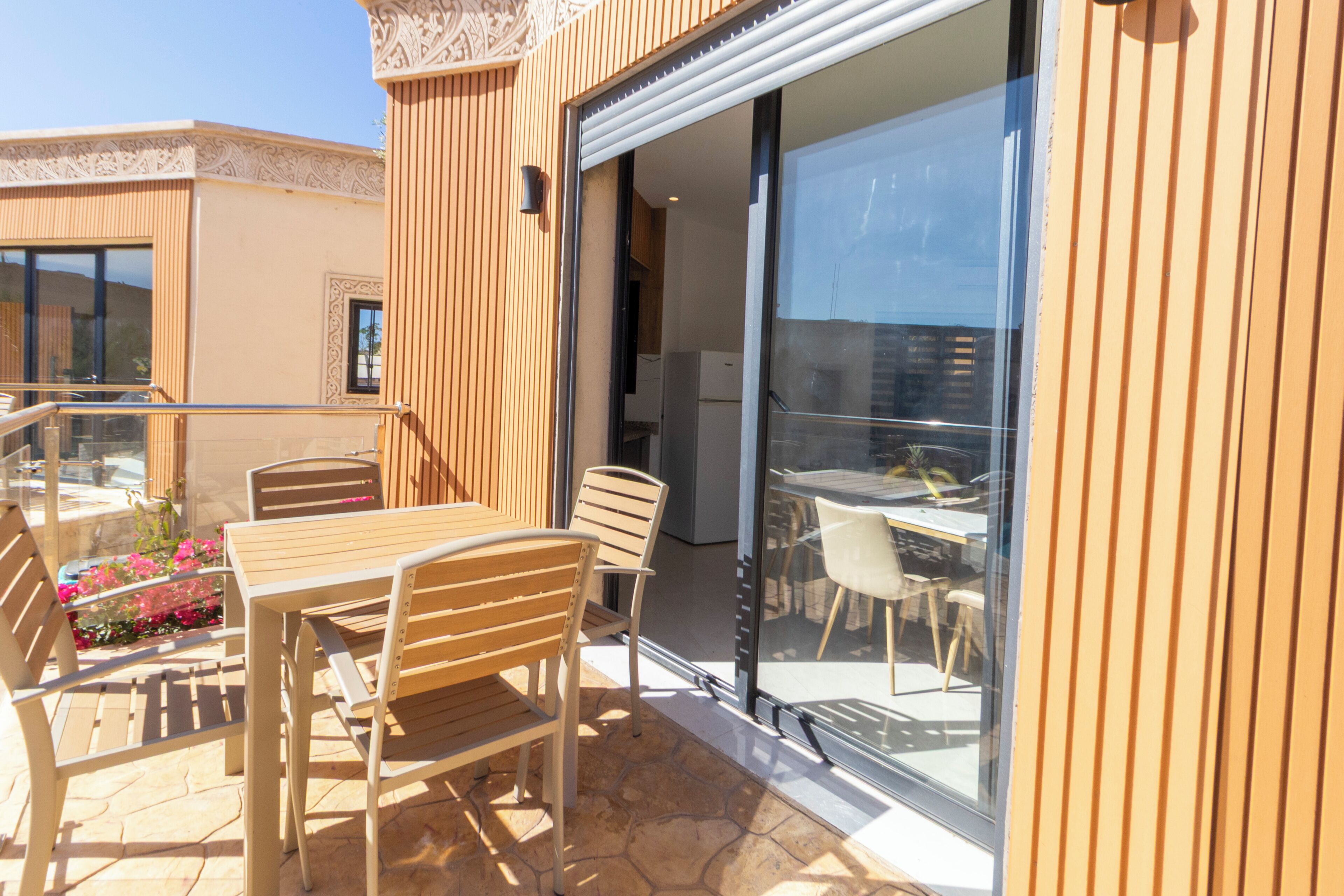 Une terrasse extérieure avec une table et des chaises en bois, une porte vitrée donnant sur un coin repas et un sol en pierre.