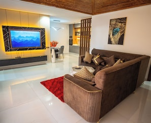 A modern living room with a brown sofa, a red rug, a flat-screen TV, and a kitchen in the background. - rose garden resort marrakech (Marrakech)