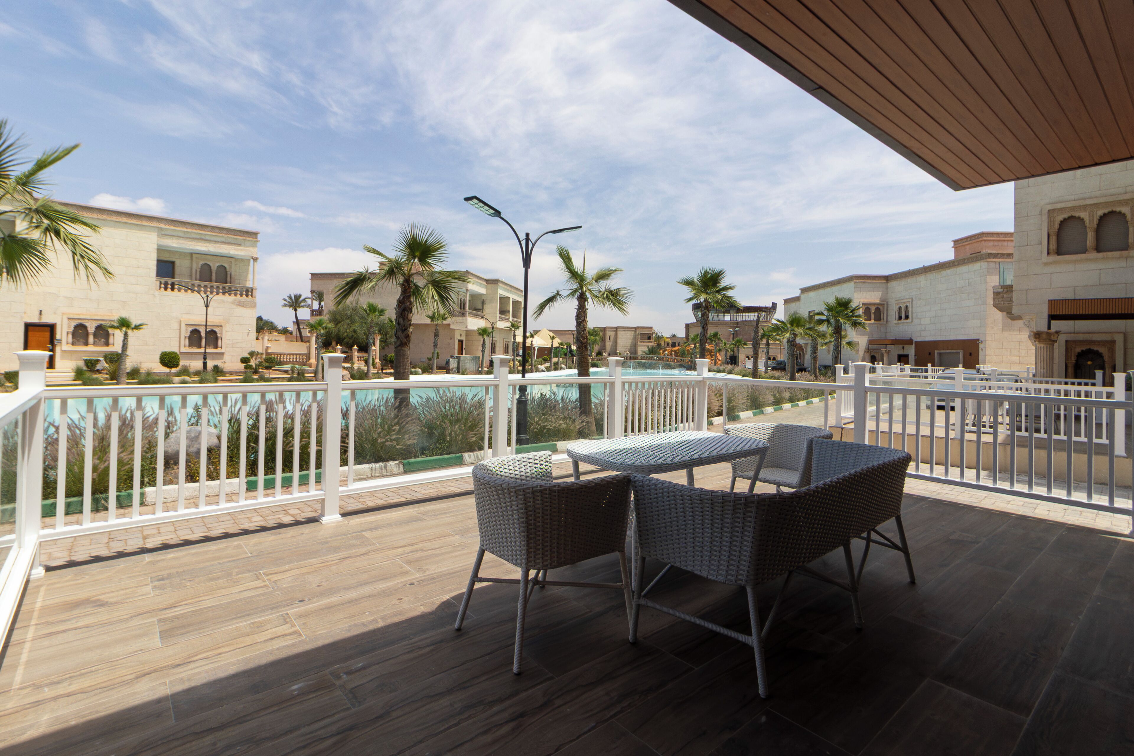 Une terrasse en bois, agrémentée de meubles en osier, donnant sur une piscine, le tout entouré de palmiers et de bâtiments à l’architecture traditionnelle.
