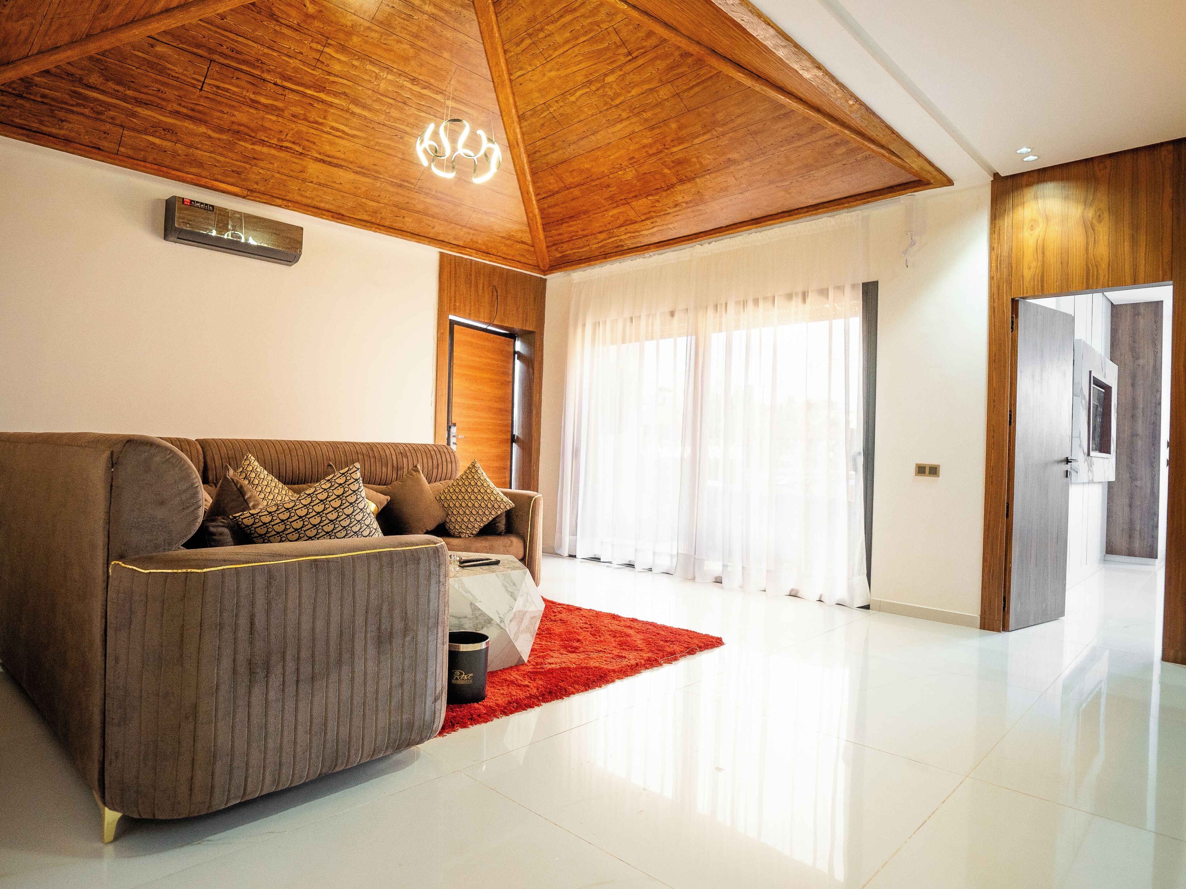 Una sala de estar moderna con techo de madera, un sofá con cojines estampados, una alfombra roja y un ventanal con cortinas traslúcidas.