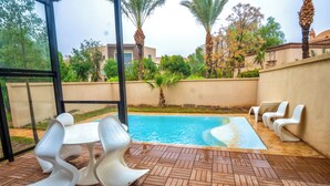 A swimming pool with a tiled deck, surrounded by palm trees and a building in the background. - rose garden resort marrakech (Marrakech)