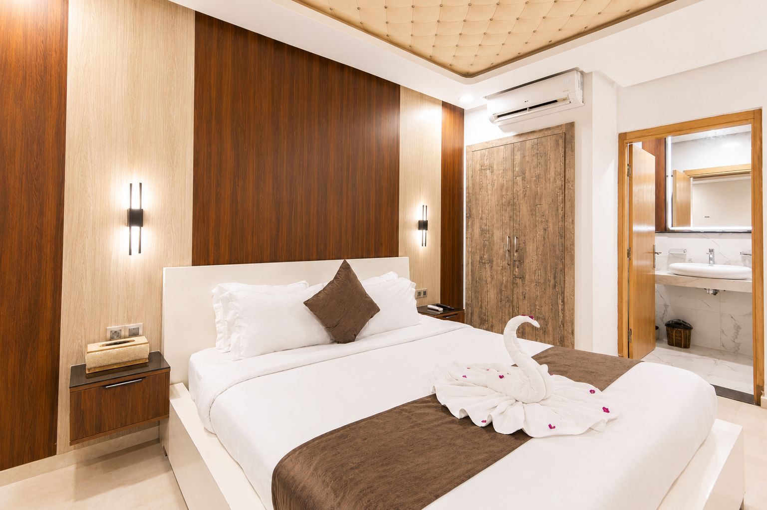 A modern hotel room with a large bed, wooden panel walls, a built-in wardrobe, a vanity with a mirror, and an air conditioning unit.