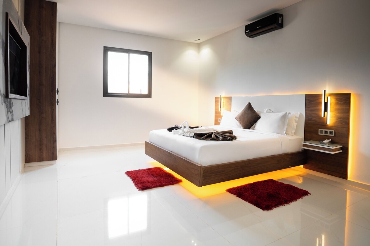 Un dormitorio moderno con una cama grande, una ventana, aire acondicionado de pared y una mesita de noche con un teléfono.