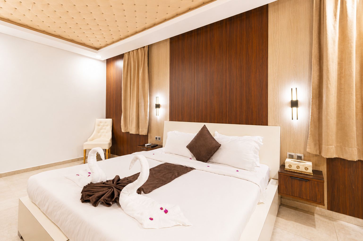 A hotel room with a large bed, a bedside table, a chair, and a wooden wall panel.