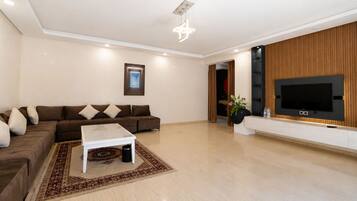 A modern living room with a sectional sofa, a coffee table, a flat-screen TV mounted on a wooden wall, and a decorative light fixture.