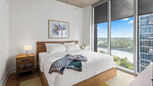 Ein Schlafzimmer mit einem großen Bett, einem Nachttisch, einem Wandbild und Blick auf eine Stadtkulisse durch große Fenster.
