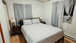 Ein Schlafzimmer mit Holzboden, einem Bett, einem Nachttisch, einer Lampe und einem Fenster mit Vorhängen.