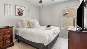 Ein Schlafzimmer mit einem Bett, einem Deckenventilator, einem Schminktisch, einem Fernseher und einem Fenster mit Ziegelmuster.