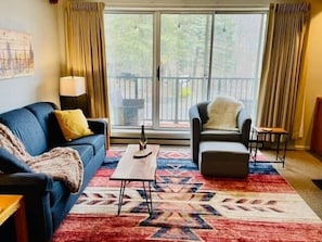 Ein Wohnzimmer mit Sofa, Sessel, Couchtisch und einem großen Fenster mit Vorhängen.