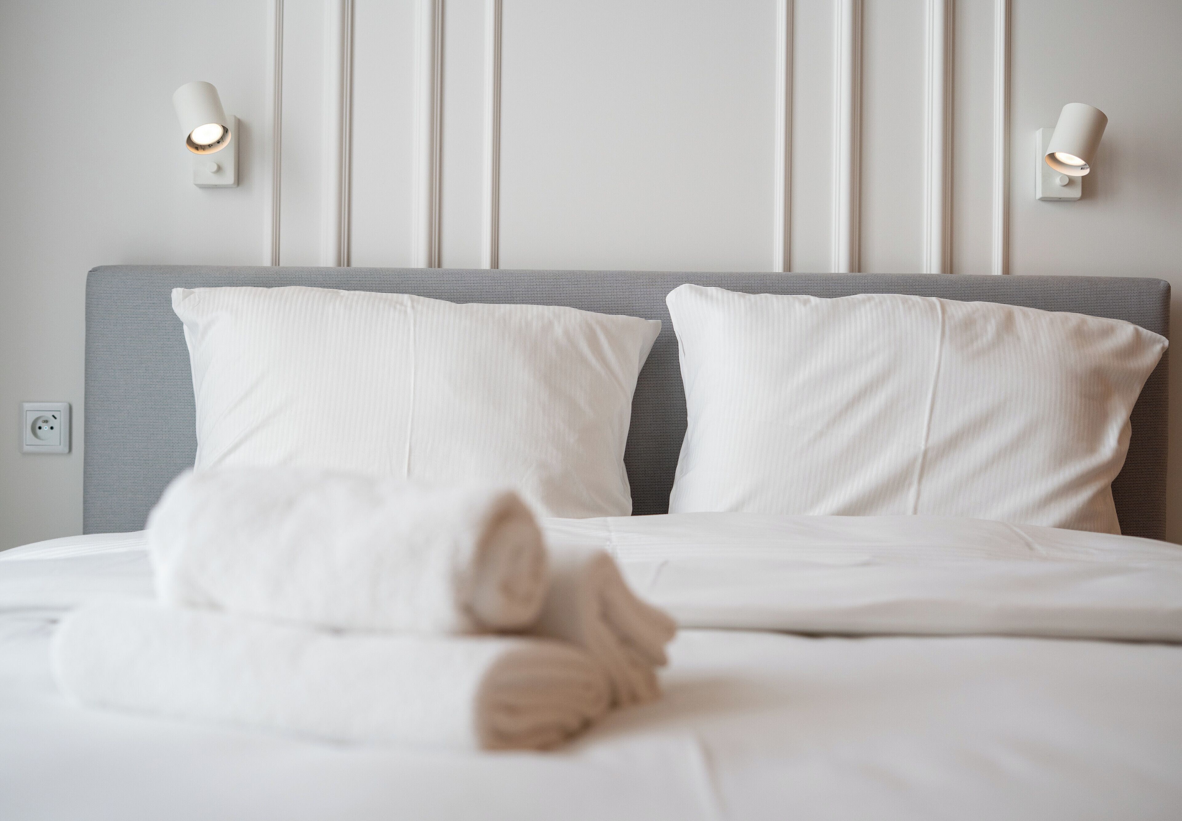 A neatly made bed with white linens and pillows, a gray headboard, and two wall-mounted light fixtures.