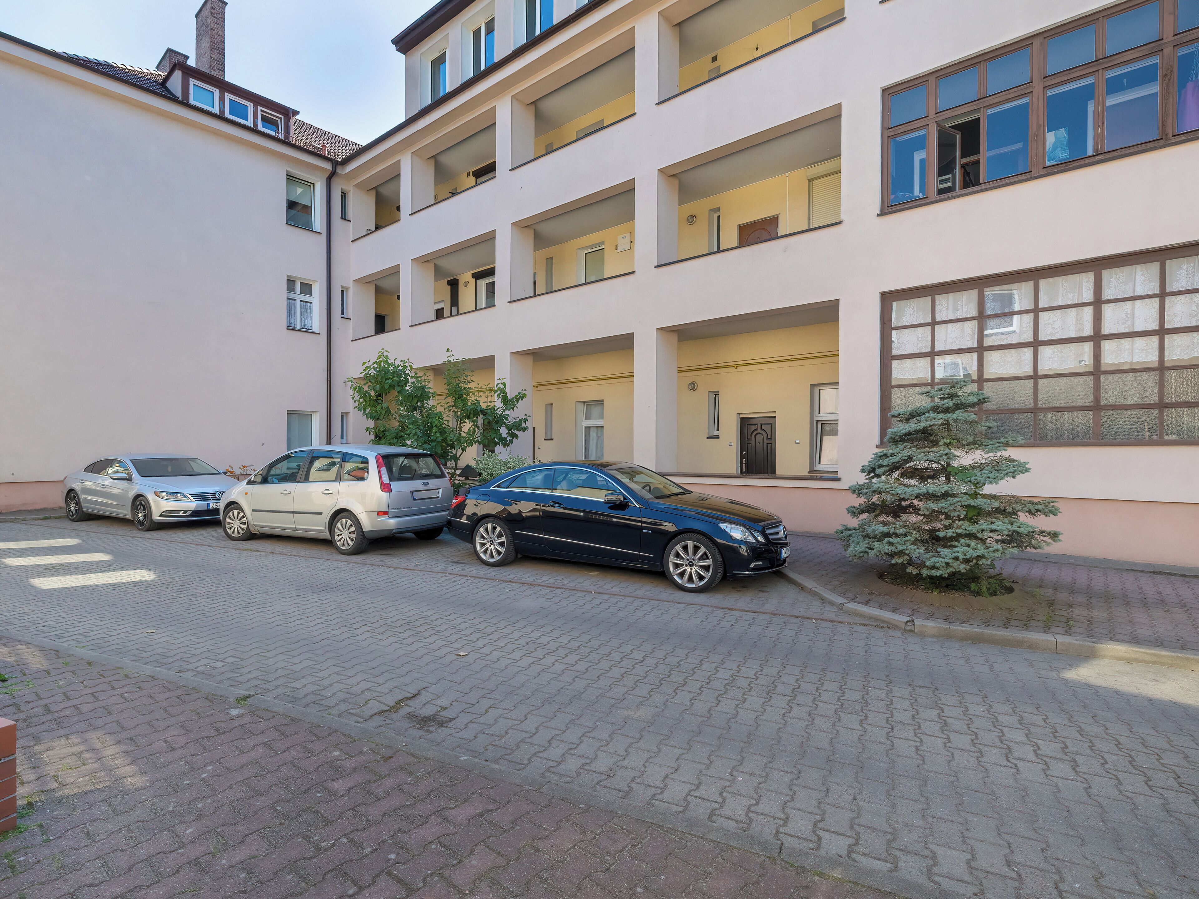 A multi-story building with a brick-paved street, parked cars, and a small tree.