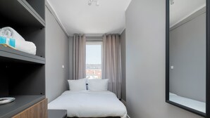 Una camera d'albergo piccola con un letto, una cassettiera in legno, uno specchio e una finestra con tende.