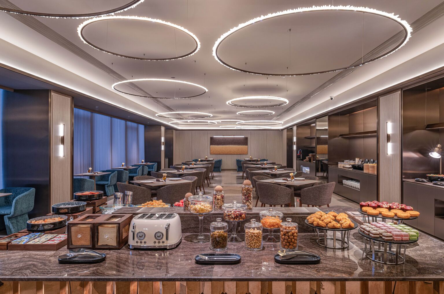 Un comedor de hotel con varias mesas, un bufé con diferentes opciones de comida y un diseño de techo moderno.