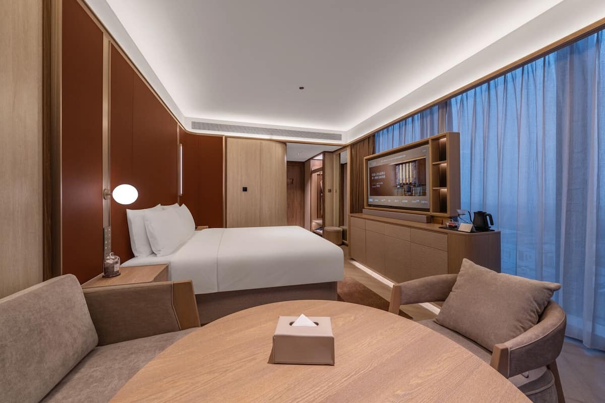 Habitación de hotel con una cama, un escritorio con lámpara, televisión y vistas a la ciudad a través de amplias ventanas.
